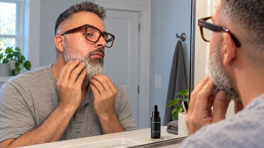 A man wearing glasses looks into a bathroom mirror and massages beard oil through his grey-flecked beard, with a bottle of Beardsage beard oil on the counter beside him.